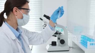 Koruyucu maske takan dişi araştırmacı ve mavi eldiven test tüpünde sıvıyı inceliyor, laboratuvarda mikroskop ve laboratuvar ekipmanlarının yanında oturuyor. Tıp, sağlık ve bilim konsepti.