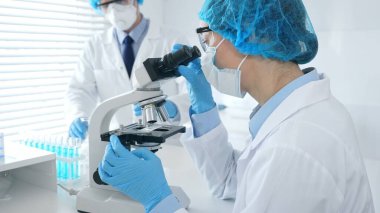 Mavi koruyucu giysiler giyen kadın bilim adamı mikroskop altında numuneleri incelerken, laboratuvar asistanı da arka planda cam parçalarıyla ilgileniyor. Tıp ve bilim..