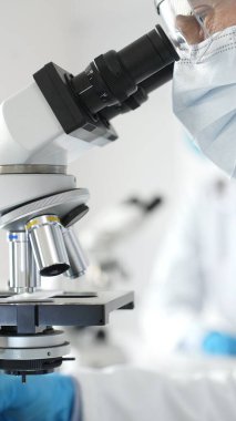 Modern laboratuvar ortamında mavi güvenlik teçhizatı takmış mikroskopla örnek inceleyen erkek araştırmacıya yakın durun. Tıp, sağlık ve bilim konsepti.