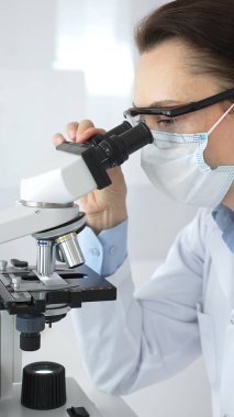 Koruyucu maskeli ve gözlüklü bir kadın bilim adamı modern bir laboratuvarda araştırma yaparken bir örneği mikroskop altında dikkatlice inceliyor. Tıp ve bilim kavramı.