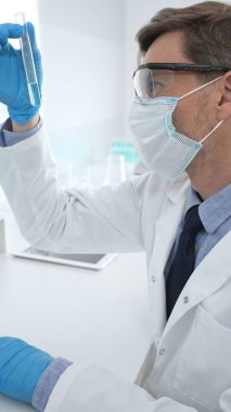 Koruyucu giysi giyen bilim araştırmacısı modern laboratuvar çalışma alanında mavi sıvı numunesini test tüpünden inceliyor. Tıp, sağlık ve bilim konsepti
