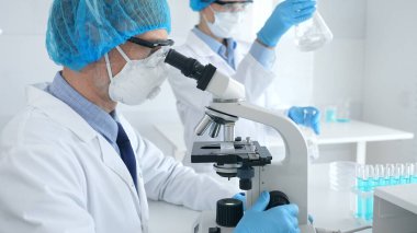 Koruyucu ekipmanlı maskeli erkek bilim adamı mikroskop altında numune incelerken laboratuvar asistanı arka planda cam eşyalar ile ilgileniyor. Tıp, bilim konsepti.
