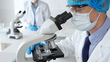Koruyucu maskeli, şapkalı ve eldivenli erkek bilim adamı steril laboratuvarda mikroskop altında örnekler üzerinde çalışıyor. Tıp, sağlık ve bilim kavramı.