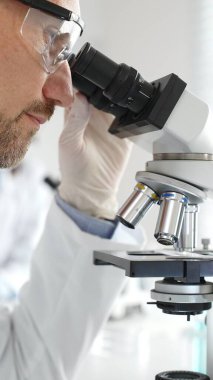 Erkek araştırmacının mikroskopla incelediği örnek, steril laboratuvar ortamında güvenlik gözlüğü takıyor, bilimsel cam eşyalar ile çevrili. Tıp, sağlık ve bilim konsepti.
