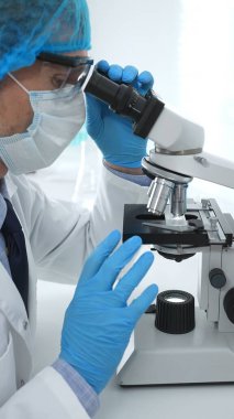 Koruyucu maske takan kıdemli erkek bilim adamı laboratuarda mikroskop kullanarak tıbbi araştırma ve analiz yapıyor. Tıp, sağlık ve bilim konsepti.
