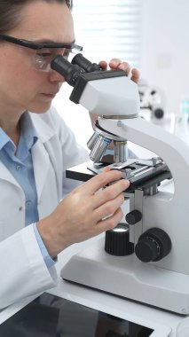 Laboratuvar önlüğü ve koruyucu gözlüklü kadın bilim adamı modern bir laboratuvarda mikroskop altında test örnekleri inceliyor. Tıp, sağlık ve bilim konsepti
