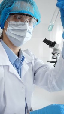 Steril laboratuvar ortamında tıbbi araştırma sırasında mikroskop yakınında mavi sıvı numunesini inceleyen koruyucu ekipman giyen bilim adamları. Tıp, sağlık ve bilim konsepti