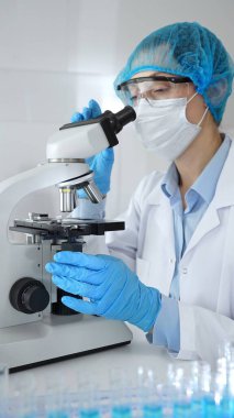 Bilinmeyen bilim adamı mavi güvenlik donanımı takıyor. Araştırma için mikroskop kullanıyor. Erkek bir meslektaşı arka planda çalışıyor. Tıp, sağlık ve bilim konsepti.