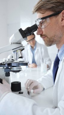 Koruyucu gözlük takan bilim adamı mikroskop altında araştırma ve analiz yapıyor. Arka planda test tüpleri ile modern bir laboratuvarda. Tıp, sağlık ve bilim konsepti