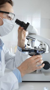 Maskeli profesyonel kadın bilim adamı mikroskop ile numune inceleyerek steril laboratuvar ortamında meslektaşlarıyla birlikte araştırma yapıyor. Tıp, sağlık ve bilim konsepti.