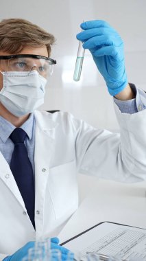 Koruyucu giysiler içindeki erkek bilim adamı modern bir laboratuvar ortamında bir pipetle turkuaz sıvıdan oluşan bir test tüpünü analiz ediyor. Tıp ve bilim konsepti.