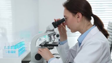Laboratuvar önlüğü ve koruyucu gözlüklü kadın bilim adamı modern bir laboratuvarda mikroskop altında test örnekleri inceliyor. Tıp, sağlık ve bilim konsepti