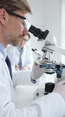 Koruyucu gözlük takan bilim adamı mikroskop altında araştırma ve analiz yapıyor. Arka planda test tüpleri ile modern bir laboratuvarda. Tıp, sağlık ve bilim konsepti