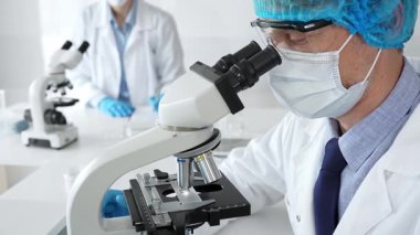 Koruyucu maske, şapka ve eldiven takan kıdemli erkek bilim adamı modern bir laboratuvarda mikroskop altında örnekleri inceliyor. Tıp, sağlık ve bilim konsepti.