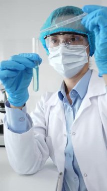 Maske takan kadın bilim adamı, mavi başlık ve eldivenler laboratuardaki pipeti kullanarak test tüpüne sıvı ekliyor. Tıp ve bilim konsepti.
