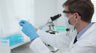 Laboratuvar önlüklü, koruyucu gözlüklü ve mavi eldivenli maskeli erkek bilim adamı modern laboratuardaki test tüplerine mikroskop ve test tüpleri rafı koyuyor. Tıp, sağlık ve bilim konsepti.