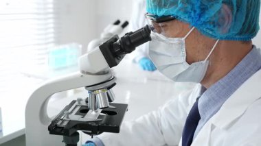 Koruyucu maske, şapka ve eldiven takan kıdemli erkek bilim adamı modern bir laboratuvarda mikroskop altında örnekleri inceliyor. Tıp, sağlık ve bilim konsepti.