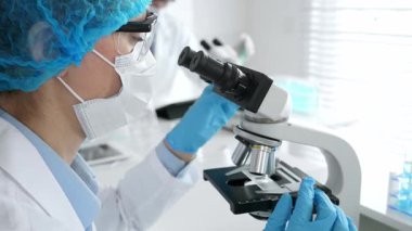 Maske takan ve mavi güvenlik donanımı takan kadın bir bilim adamı araştırma için mikroskop kullanırken erkek bir meslektaşı arka planda çalışıyor. Tıp, sağlık ve bilim konsepti.