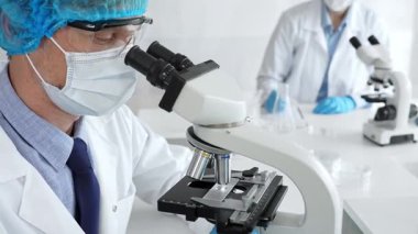 Koruyucu maske, şapka ve eldiven takan kıdemli erkek bilim adamı modern bir laboratuvarda mikroskop altında örnekleri inceliyor. Tıp, sağlık ve bilim konsepti.