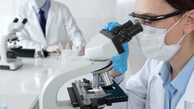 Maskeli profesyonel kadın bilim adamı mikroskop ile numune inceleyerek steril laboratuvar ortamında meslektaşlarıyla birlikte araştırma yapıyor. Tıp, sağlık ve bilim konsepti.