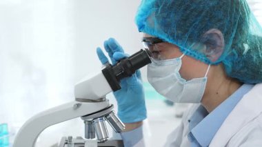 Koruyucu maske takan, gözlük takan ve mikroskop kullanarak laboratuvarda bilimsel araştırma yapan kadın bilim adamı. Tıp, sağlık ve bilim konsepti