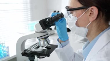 Laboratuvarda mikroskop kullanarak laboratuvar önlüğü ve güvenlik gözlüğü takan kadın bilim adamı. Arka planda deney tüpleri ile bilimsel araştırma yapıyor. Tıp, sağlık ve bilim konsepti