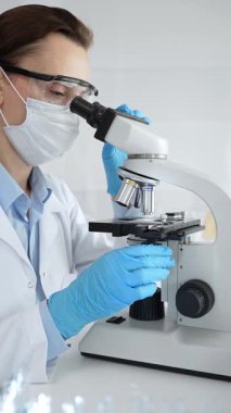 Kadın araştırmacı mikroskop ile örnek analiz ediyor, koruyucu teçhizat takılı, steril araştırma ortamında gelişmiş laboratuvar ekipmanlarının yanında çalışan bir meslektaşı. Tıp, sağlık ve bilim konsepti