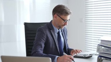 Takım elbiseli ve kravatlı, siyah gözlüklü, olgun girişimci, masada oturuyor, notlar yazıyor ve modern, parlak ofiste hesap makinesi kullanıyor. Ticarette denetim ve vergiler.