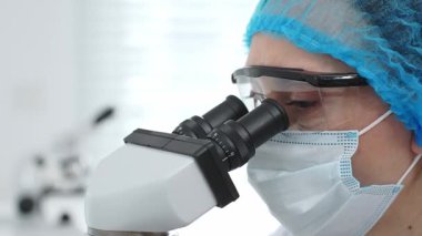 Mikroskop kullanan, maske, gözlük ve eldiven takan bir bilim adamının laboratuvar ortamında araştırma yapmasını. Tıp, sağlık ve bilim konsepti