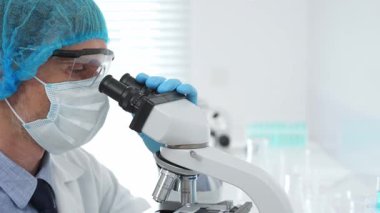 Kıdemli bilim adamı mikroskopla numune inceliyor, steril laboratuvar ortamında tıbbi araştırma yaparken koruyucu giysiler giyiyor. Tıp, sağlık ve bilim konsepti
