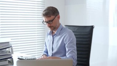 Mavi gömlekli ve gözlüklü profesyonel bir muhasebeci ofis ortamında mali raporları incelerken fatura hesaplıyor ve faturaları yönetiyor. İş dünyasında denetim ve vergi teması.