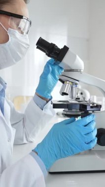 Kadın araştırmacı mikroskop ile örnek analiz ediyor, koruyucu teçhizat takılı, steril araştırma ortamında gelişmiş laboratuvar ekipmanlarının yanında çalışan bir meslektaşı. Tıp, sağlık ve bilim konsepti