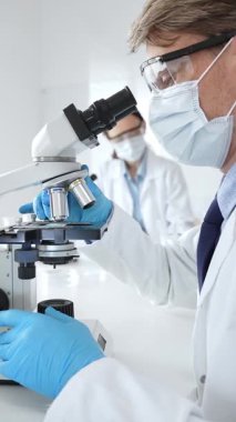 Mikroskop kullanarak maske takan ve eldiven takan bilim adamlarına yakın çekim, modern laboratuvarda araştırmalar yürütmek ve arka planda çalışmak. Tıp, sağlık ve bilim konsepti