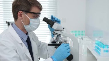 Laboratuvar araştırmacısı mavi sıvı örneklerini analiz ediyor, mikroskop altında detaylı bilimsel araştırma yaparken koruyucu giysi giyiyor. Tıp, sağlık ve bilim konsepti