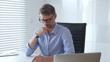 Mavi gösterişli gömlek ve gözlük giyen işadamı, parlak, modern bir ofiste çalışırken düşünüyor ve konsantre oluyor, profesyonelliğini gösteriyor. İş adamları konsepti