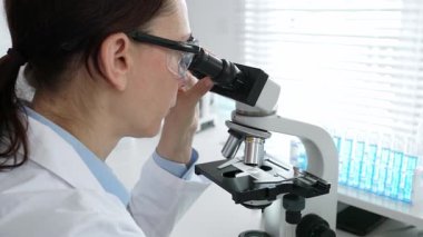 Laboratuvarda mikroskop kullanarak laboratuvar önlüğü ve güvenlik gözlüğü takan kadın bilim adamı. Arka planda deney tüpleri ile bilimsel araştırma yapıyor. Tıp, sağlık ve bilim konsepti