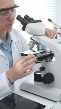 Laboratuvar önlüğü ve koruyucu gözlüklü kadın bilim adamı modern bir laboratuvarda mikroskop altında test örnekleri inceliyor. Tıp, sağlık ve bilim konsepti