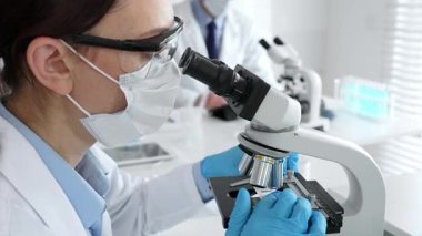 Maskeli profesyonel kadın bilim adamı mikroskop ile numune inceleyerek steril laboratuvar ortamında meslektaşlarıyla birlikte araştırma yapıyor. Tıp, sağlık ve bilim konsepti.