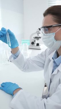 Mavi koruyucu eldivenli maskeli kadın araştırmacı test tüpündeki temiz sıvıyı inceliyor, mikroskop ve laboratuvar ekipmanlarının yanında oturuyor. Tıp, sağlık ve bilim konsepti.