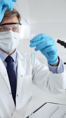 Maske, mavi başlık ve eldiven takan erkek bilim adamı test tüpüne sıvı ekliyor. Laboratuvarda pipet kullanarak tıbbi araştırma yapıyor. Tıp ve bilim konsepti.