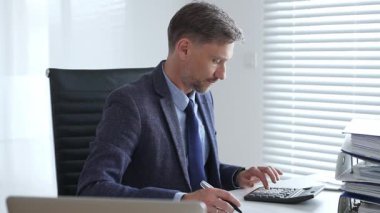 Takım elbiseli ve kravatlı bir işadamı modern ofiste mali belgelerle uğraşırken masada oturuyor ve hesap makinesi kullanıyor. İş dünyasında denetim ve vergi teması