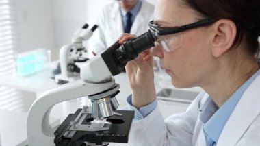 Koruyucu gözlük takan kadın bilim adamı mikroskop kullanarak modern laboratuvarda deney tüpleri ve arka planda erkek bilim adamı ile araştırma yapıyor. Tıp, sağlık ve bilim konsepti