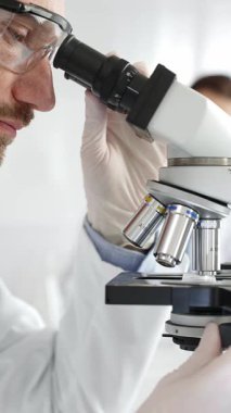 Erkek araştırmacının mikroskopla incelediği örnekler steril laboratuvar ortamında güvenlik gözlüğü takıyor. Tıp, sağlık ve bilim konsepti.