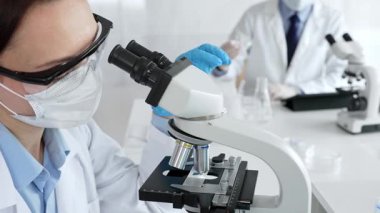 Maskeli profesyonel kadın bilim adamı mikroskop ile numune inceleyerek steril laboratuvar ortamında meslektaşlarıyla birlikte araştırma yapıyor. Tıp, sağlık ve bilim konsepti.