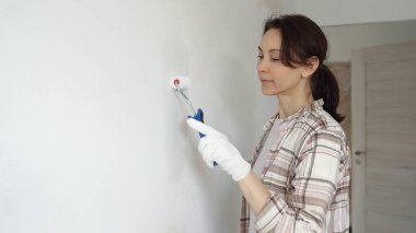 Koruyucu eldivenler giyen kadın, beyaz duvarı küçük boya rulosuyla boyuyor, iç dekorasyonu yeniliyor ve ev yenileme projesinde odayı temizliyor..