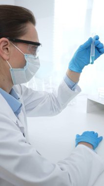 Laboratuvar önlüğü giyen ve koruyucu gözlük takan kadın bilim adamı modern laboratuarda mikroskop ve test tüpleri askısı ile test tüpünü temiz sıvıyla analiz ediyor. Tıp, sağlık ve bilim konsepti