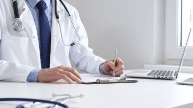 Erkek doktor reçete yazıyor ve bir ofis ortamındaki bir panoya tıbbi kayıtları dolduruyor. Sağlık ve hasta bakımının temel yönlerini gösteriyor. İlaç ve sağlık hizmetleri.