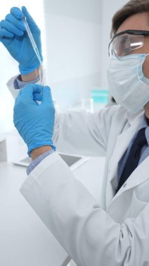 Laboratuvar önlüklü, koruyucu gözlüklü ve mavi eldivenli maskeli erkek bilim adamı modern laboratuardaki test tüplerine mikroskop ve test tüpleri rafı koyuyor. Tıp, sağlık ve bilim konsepti.