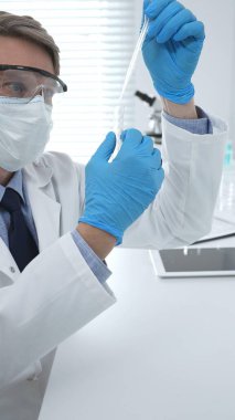 Laboratuvar önlüklü, koruyucu gözlüklü ve mavi eldivenli maskeli erkek bilim adamı modern laboratuardaki test tüplerine mikroskop ve test tüpleri rafı koyuyor. Tıp, sağlık ve bilim konsepti.