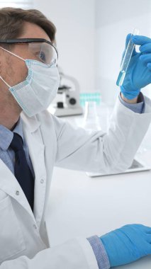 Koruyucu giysi giyen bilim araştırmacısı modern laboratuvar çalışma alanında mavi sıvı numunesini test tüpünden inceliyor. Tıp, sağlık ve bilim konsepti
